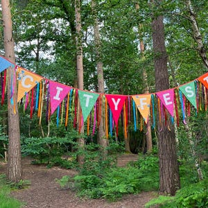 May include: A colorful banner with the text "CITY FEST" strung between trees in a forest. The banner is made of triangular flags in various colors, including blue, orange, pink, and green, with colorful ribbons hanging down.