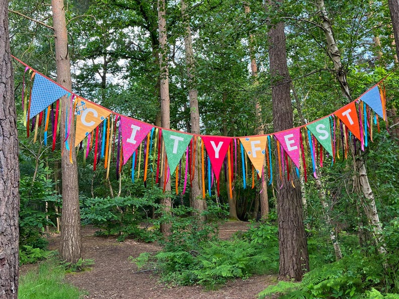 May include: A colorful banner with the text "CITY FEST" strung between trees in a forest. The banner is made of triangular flags in various colors, including blue, orange, pink, and green, with colorful ribbons hanging down.