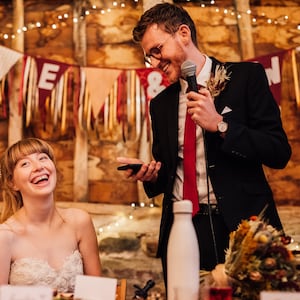 May include: A candid moment at a wedding reception. A smiling person in a strapless white dress sits at a table, while a person in a black suit with a red tie speaks into a microphone. Decorative banners and string lights adorn the background.