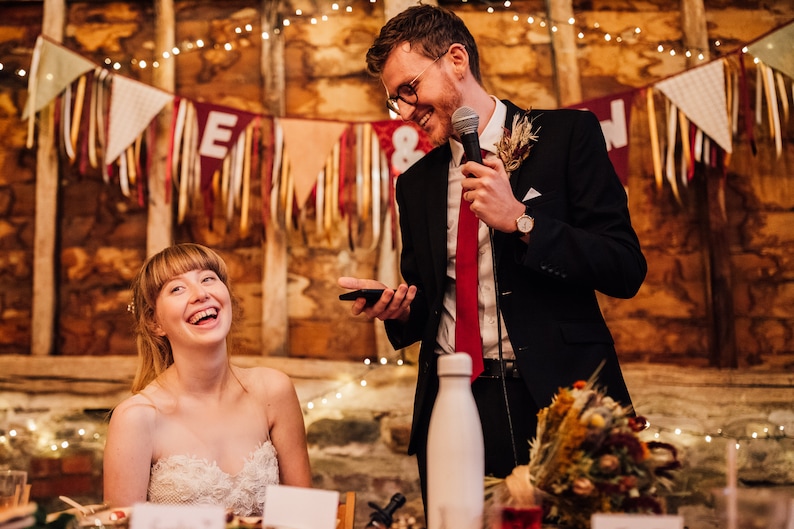 May include: A candid moment at a wedding reception. A smiling person in a strapless white dress sits at a table, while a person in a black suit with a red tie speaks into a microphone. Decorative banners and string lights adorn the background.
