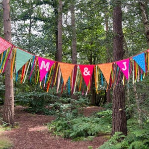 May include: A colorful banner with pennant flags strung between trees in a forest. The flags are various colors, including pink, green, orange, and red, with the letters "M & J" displayed. Ribbons of different colors hang from the flags.