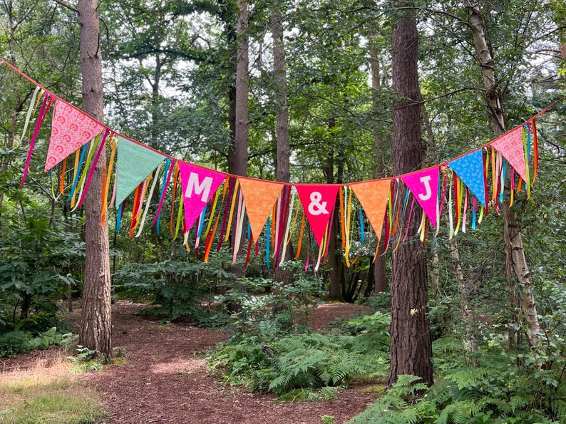 May include: A colorful banner with pennant flags strung between trees in a forest. The flags are various colors, including pink, green, orange, and red, with the letters "M & J" displayed. Ribbons of different colors hang from the flags.