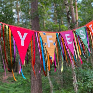 May include: A colorful banner with the word "Y FEST" spelled out on triangular flags. The flags are red, orange, green, and pink, with white letters. Long, colorful ribbons hang from the bottom of the flags. The banner is set against a backdrop of trees.