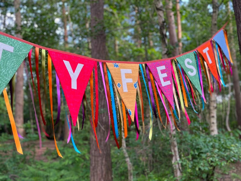 May include: A colorful banner with the word "Y FEST" spelled out on triangular flags. The flags are red, orange, green, and pink, with white letters. Long, colorful ribbons hang from the bottom of the flags. The banner is set against a backdrop of trees.