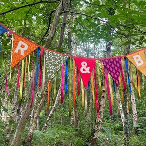 May include: A colorful banner with pennant flags and ribbons hangs in a forest. The flags display letters and patterns, including the letters "R & B". The ribbons are in various colors, adding to the festive look.