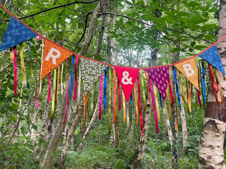 May include: A colorful banner with pennant flags and ribbons hangs in a forest. The flags display letters and patterns, including the letters "R & B". The ribbons are in various colors, adding to the festive look.