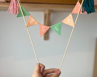 May include: A felt banner with the word "It's a" in white lettering on peach and green triangular flags. The banner is attached to a wooden stick with pink and green yarn tassels at each end.