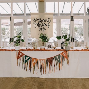 May include: A wedding reception table is decorated with a white tablecloth, orange runner, and a banner that reads "You Were the Obvious One." A banner with the word "TEAM" hangs in front. Green plants and candles are placed on the table.