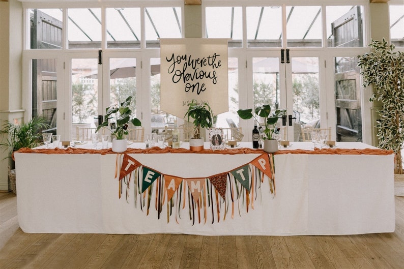 May include: A wedding reception table is decorated with a white tablecloth, orange runner, and a banner that reads "You Were the Obvious One." A banner with the word "TEAM" hangs in front. Green plants and candles are placed on the table.