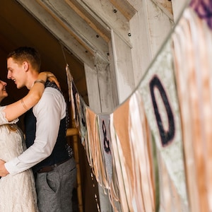 May include: A couple embraces, smiling at each other. The woman wears a lace wedding dress and veil. The man is in a vest and dress pants. A banner with letters hangs in the background, with a rustic wooden structure.