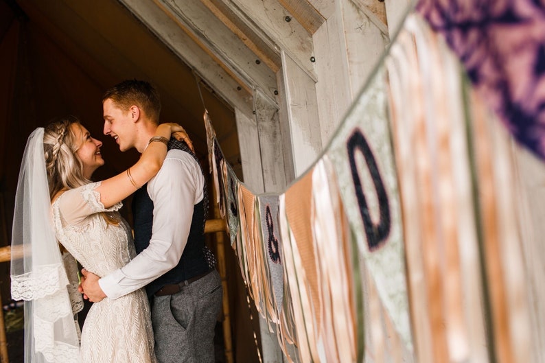 May include: A couple embraces, smiling at each other. The woman wears a lace wedding dress and veil. The man is in a vest and dress pants. A banner with letters hangs in the background, with a rustic wooden structure.