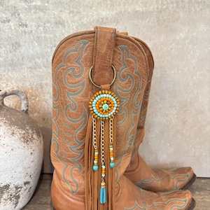May include: A tan leather cowboy boot featuring intricate teal stitching and a decorative accessory attached to the pull tab. The accessory consists of a circular beaded medallion in orange and teal, gold chains, and long tan leather fringe.