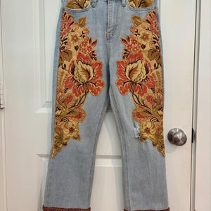 May include: Light blue denim jeans with elaborate floral embroidery in shades of red, orange, and gold. The jeans feature a distressed knee and a fringed hemline, offering a unique, bohemian aesthetic.