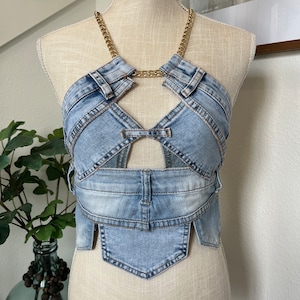 May include: A light blue denim crop top with a gold chain halter neckline and a unique, layered design. The top has a cut-out detail at the center front and a pointed hemline.
