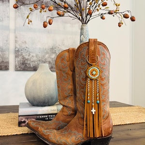 May include: A pair of brown leather cowboy boots with turquoise and brown embroidered details. The boots are decorated with a beaded and fringed boot charm. A decorative fall arrangement is in the background.