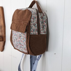 May include: A brown and white floral patterned backpack with a brown fleece flap and a brown fleece bottom. The backpack has a zipper closure and two blue adjustable straps with a white logo.