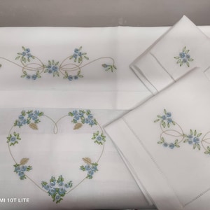 Puede incluir: Ropa de cama blanca bordada con diseños florales azules. El bordado incluye un patrón en forma de corazón y arreglos florales. El conjunto incluye una sábana y dos fundas de almohada, todas con costuras delicadas.
