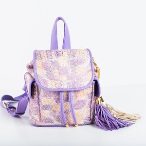 May include: A small, purple backpack with a snakeskin pattern in shades of lavender, cream, and beige. The bag has a top handle, two side pockets, and a drawstring closure. A long, purple tassel hangs from the side. The bag has a gold chain and hardware.