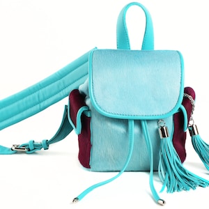 Op de afbeelding: Een kleine turquoise blauwe rugzak met een bordeauxrode suède accent en twee lange turquoise kwastjes. De rugzak heeft een bovenste handvat en een lange verstelbare band.