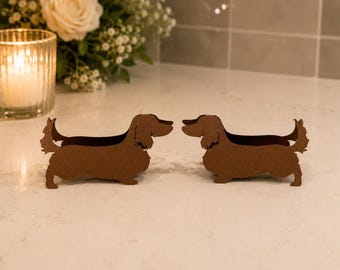 May include: Two brown, flat, laser-cut silhouettes of long-haired dachshund dogs facing each other. The dog figures are made of a solid brown material and feature detailed outlines of the ears, tails, and paws in a standing profile position.