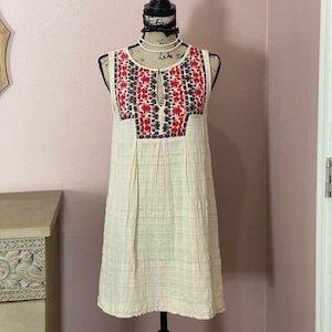 Puede incluir: Un vestido blanco de algodón sin mangas con bordados rojos y morados en el corpiño. El vestido tiene un escote redondo y una abertura de ojo de cerradura en la parte delantera.