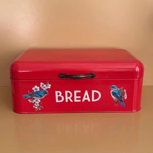 Puede incluir: Una panera de metal roja con tapa abisagrada y asa negra. La palabra "BREAD" está impresa en blanco en la parte delantera, junto con dos ilustraciones de pájaros azules y flores blancas. La panera es rectangular.