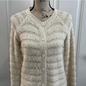 May include: A cream-colored knitted cardigan with a button-up front and long sleeves. The cardigan has a textured, fuzzy knit pattern.