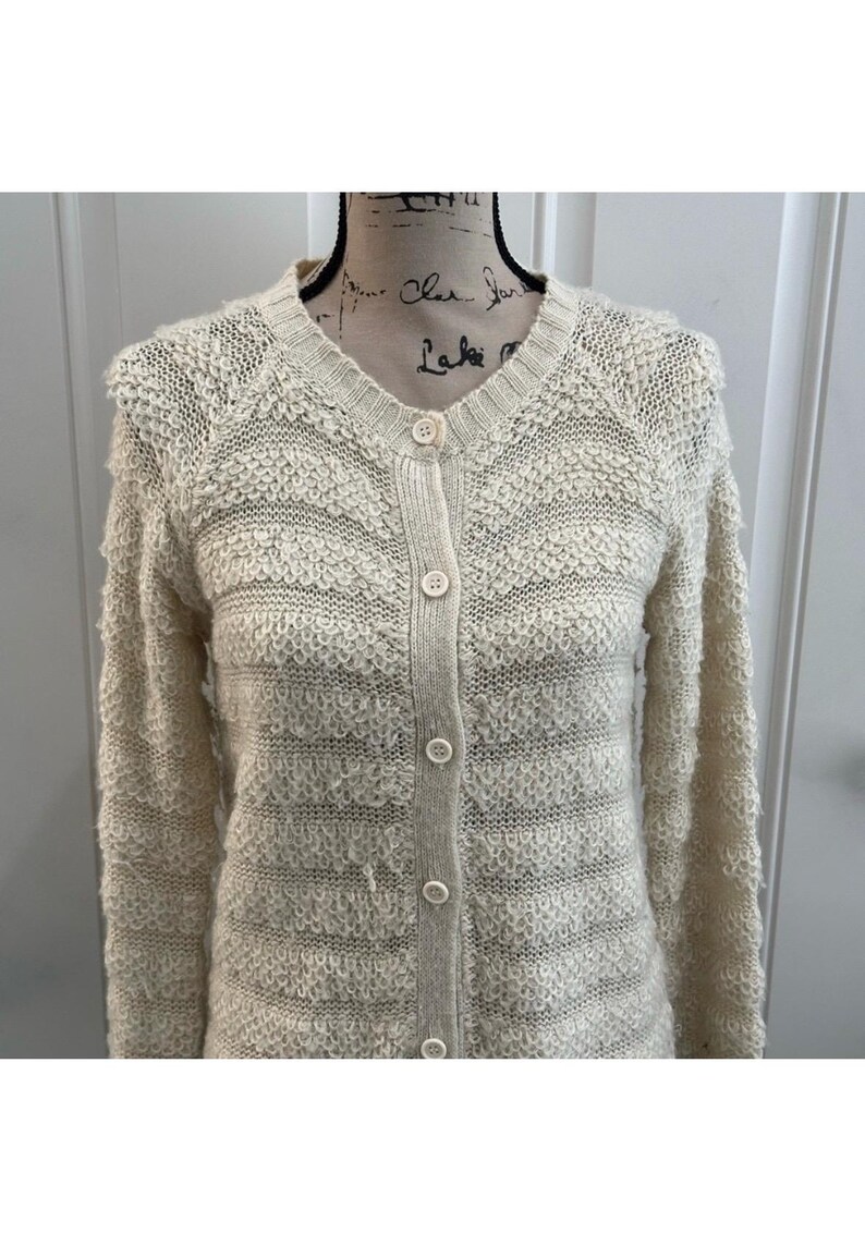May include: A cream-colored knitted cardigan with a button-up front and long sleeves. The cardigan has a textured, fuzzy knit pattern.