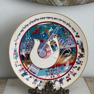 May include: Decorative ceramic plate with a gold rim, featuring a dove design and colorful abstract art. The plate has Hebrew text around the edge and the word "Abcassis" in the center. It is displayed on a decorative metal stand.