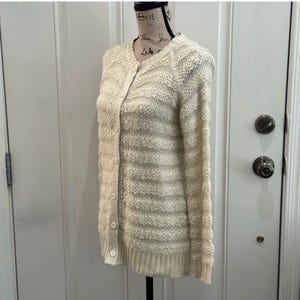 May include: A cream-colored knitted cardigan with a button-up front and long sleeves. The cardigan has a textured knit pattern and a striped design.