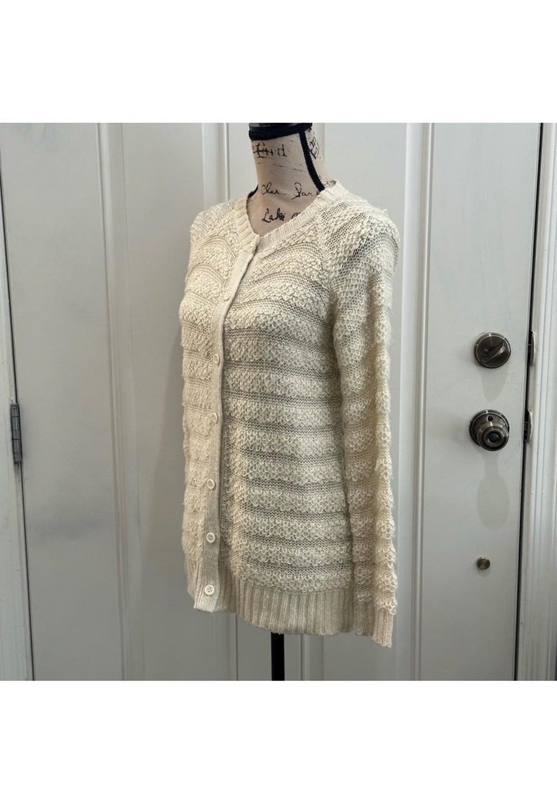 May include: A cream-colored knitted cardigan with a button-up front and long sleeves. The cardigan has a textured knit pattern and a striped design.