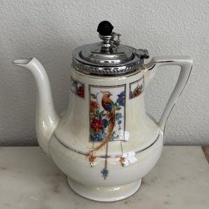 Op de afbeelding: Crèmekleurige vintage theepot met een zilveren deksel en handvat. De theepot heeft een kleurrijk vogel- en bloemmotief. De tuit en het handvat zijn zilver omlijnd. Het deksel heeft een zwarte knop.