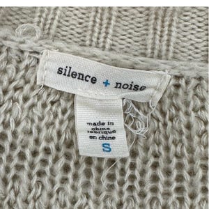 May include: A white tag with the text "silence + noise" and "made in china / fabrique en chine" and a size "S" on a beige knitted sweater.