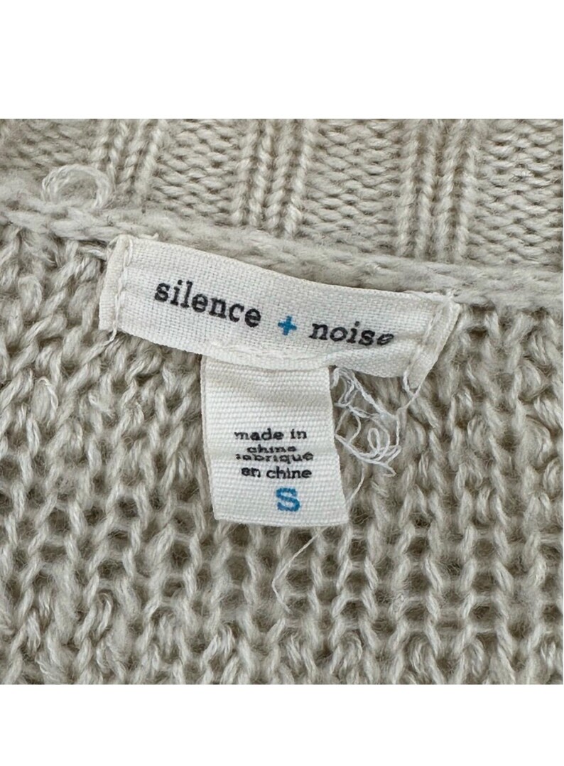 May include: A white tag with the text "silence + noise" and "made in china / fabrique en chine" and a size "S" on a beige knitted sweater.