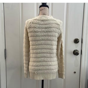 May include: A cream-colored knitted sweater with a striped pattern. The sweater is on a mannequin and has long sleeves.