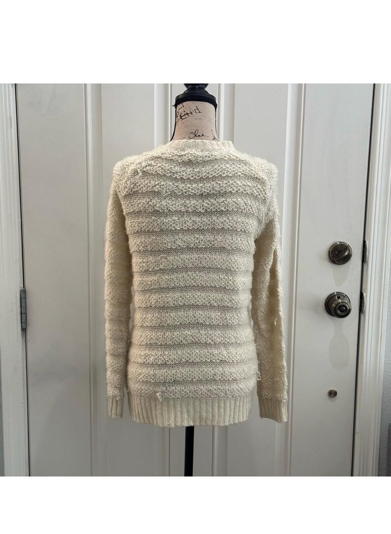 May include: A cream-colored knitted sweater with a striped pattern. The sweater is on a mannequin and has long sleeves.