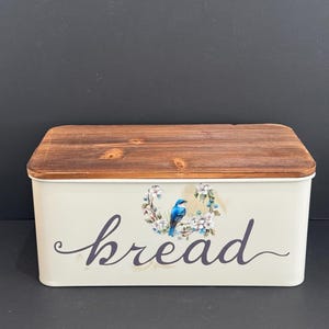 May include: A rectangular cream-colored metal bread box with a dark brown wooden lid. The front features the word bread in a dark cursive script, centered below a small illustration of a blue bird perched on a branch surrounded by white flowers.