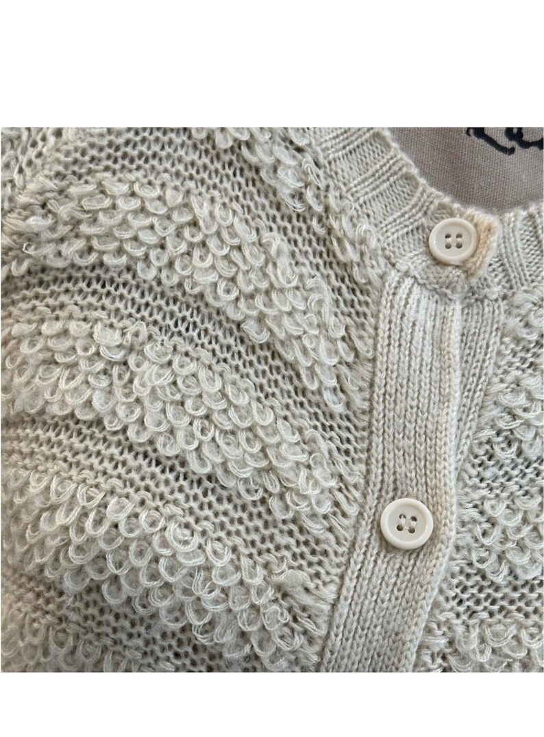 May include: A close-up of a cream-colored knitted cardigan with a textured pattern. The cardigan has a button closure and is made of a soft, fluffy material.