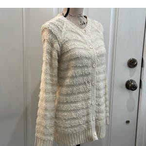 May include: A cream-colored knitted cardigan with a button-up front and a striped pattern. The cardigan has long sleeves and a relaxed fit.