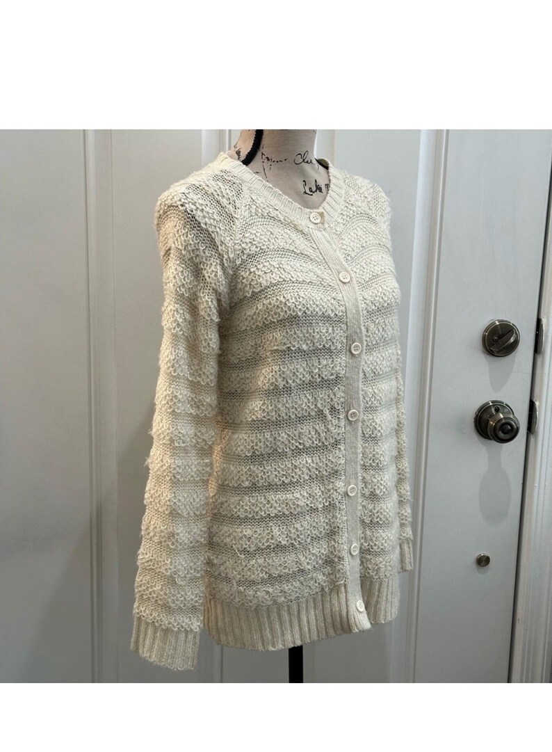 May include: A cream-colored knitted cardigan with a button-up front and a striped pattern. The cardigan has long sleeves and a relaxed fit.