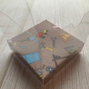 May include: A clear, transparent, plastic container with a brown paper base. The base features colorful illustrations of construction tools and vehicles, including a drill, hammer, and truck. The container is open at the top.