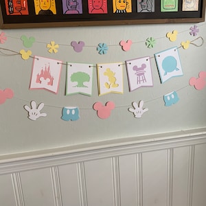 May include: A pastel-colored paper banner with Mickey Mouse and Disney-themed cutouts. The banner includes images of Mickey Mouse, the Disney castle, a tree, a ride, and Mickey Mouse's iconic gloves. The banner is strung with twine and has a light green background.