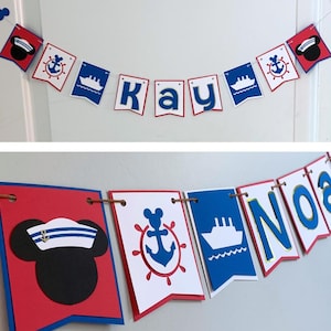 May include: A nautical-themed banner with red, white, and blue pennants. The banner spells out "Kay" and "Noah" with nautical symbols like anchors, ships, and a sailor hat. The banner is strung on a thin twine.