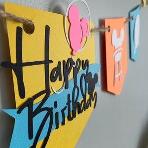 May include: A colorful banner with the words "Happy Birthday" cut out in black script. The banner features yellow, blue, and orange pennants, with a pink Mickey Mouse balloon design. The banner is strung on a natural twine.