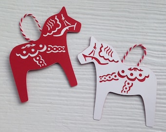 May include: Two wooden Scandinavian Dala horse ornaments, one red with white details and one white with red details. The ornaments are hanging from a white string.