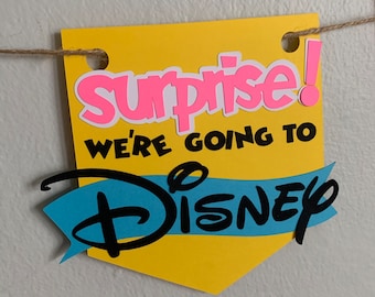 Puede incluir: Una pancarta amarilla con el texto "Surprise! We're going to Disney" en letras negras y rosas. El logotipo de Disney está en negro con una cinta azul detrás.