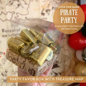 May include: A clear plastic box filled with gold chocolate nuggets, with a brown paper treasure map design on the bottom. The box is labeled "Hershey's Nuggets" and is part of a pirate party favor set.