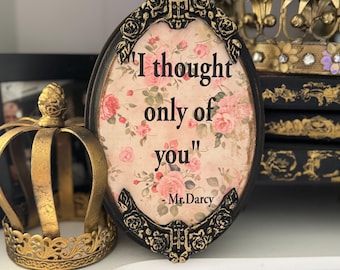 May include: A black oval frame with a gold trim and a floral background. The frame contains a quote from Mr. Darcy from Pride and Prejudice: "I thought only of you".