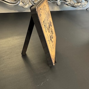 May include: A rectangular wooden sign with a black frame and a tan face featuring the text BELLADONNA, botanical illustrations of plants, and decorative scrollwork. The sign is supported by a black easel back stand for upright display.