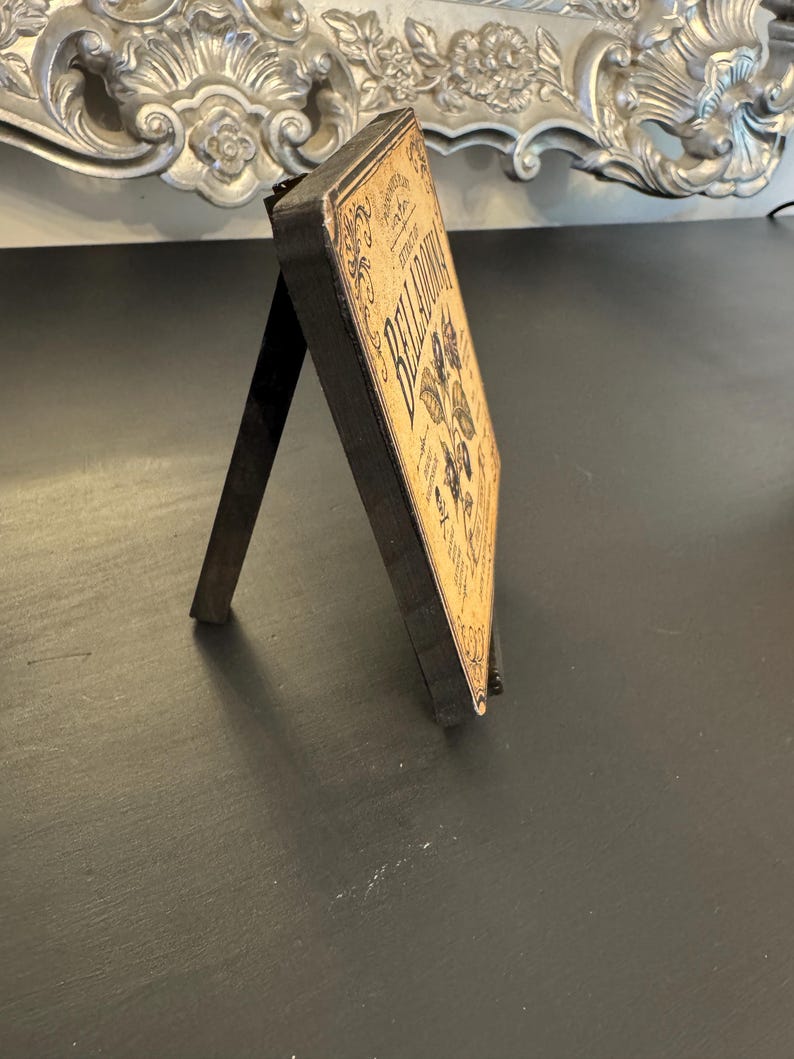 May include: A rectangular wooden sign with a black frame and a tan face featuring the text BELLADONNA, botanical illustrations of plants, and decorative scrollwork. The sign is supported by a black easel back stand for upright display.